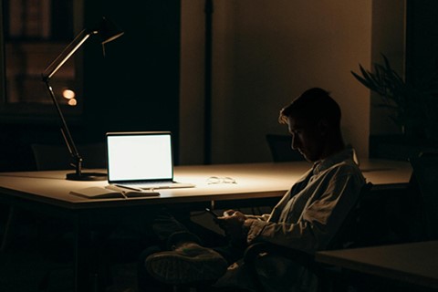 En man i en kontorsstol med en laptop på skrivbordet och en lampa som lyser i det annars mörka rummet