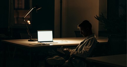 En man i en kontorsstol med en laptop på skrivbordet och en lampa som lyser i det annars mörka rummet