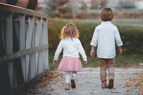 Två ljusklädda små barn syns bakifrån gående på en väg med höstlöv.